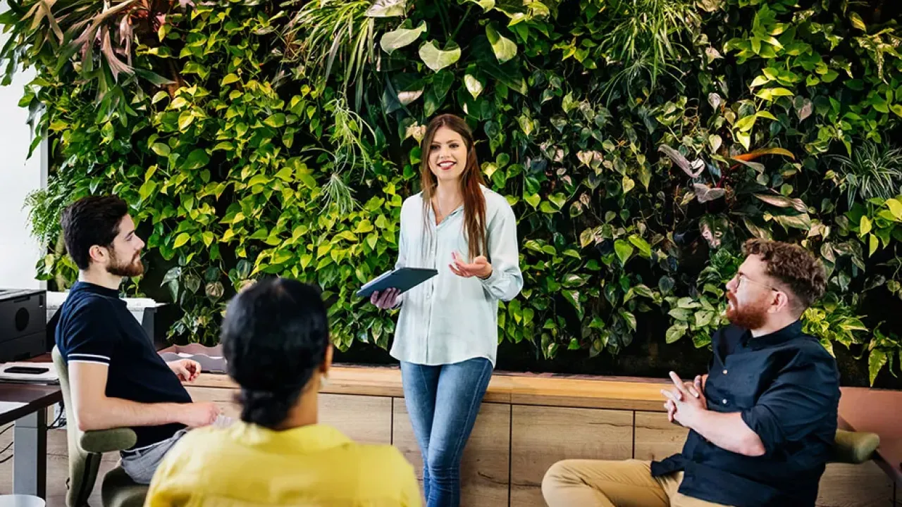 young employee in a white top stands and talks in front of a wall of green plants while coworkers listen