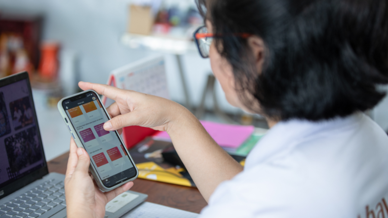 Looking over a person's shoulder, they are using the HerVenture app on a cell phone. A laptop open in front of them on a desk.