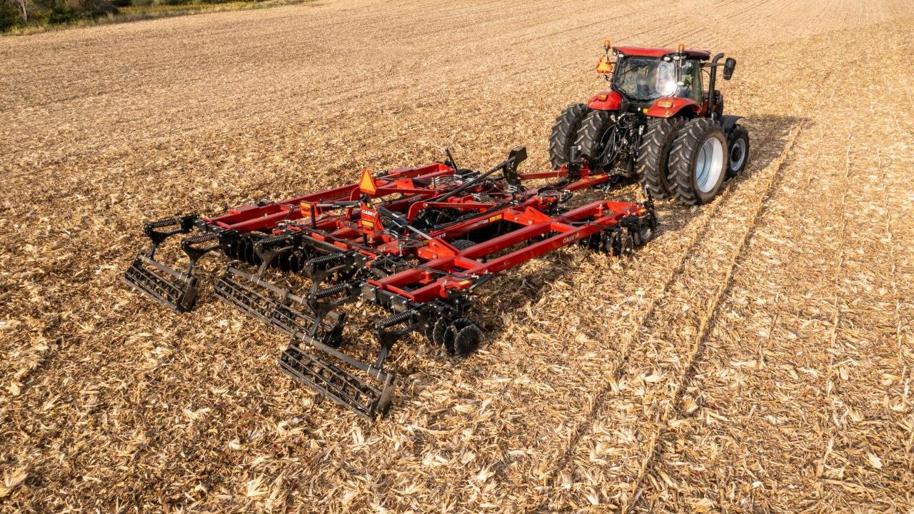 A large red tractor and attached tilling tool on a large harvested crop field.