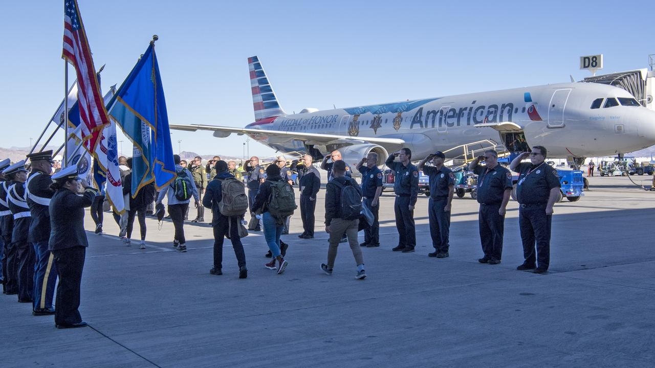 On the tarmac, lines of uniformed officers holding flags and holding salute as other pass between them. A plane behind them.