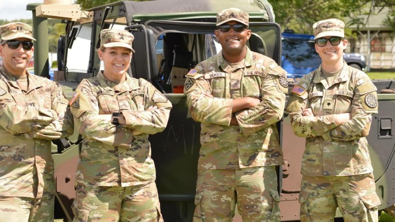 Four people posed with arms crossed in military uniforms. A military vehicle behind them.