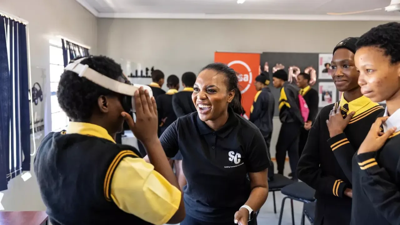 An adult smiles and assists a child with a vr headset. Others look on smiling.