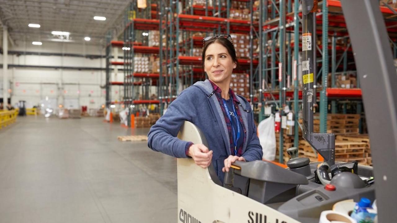 A woman in a warehouse stands in the operators seat of a forklift. She looks over her right shoulder and smiles with floor to ceiling shelves stacked with boxes behind her.