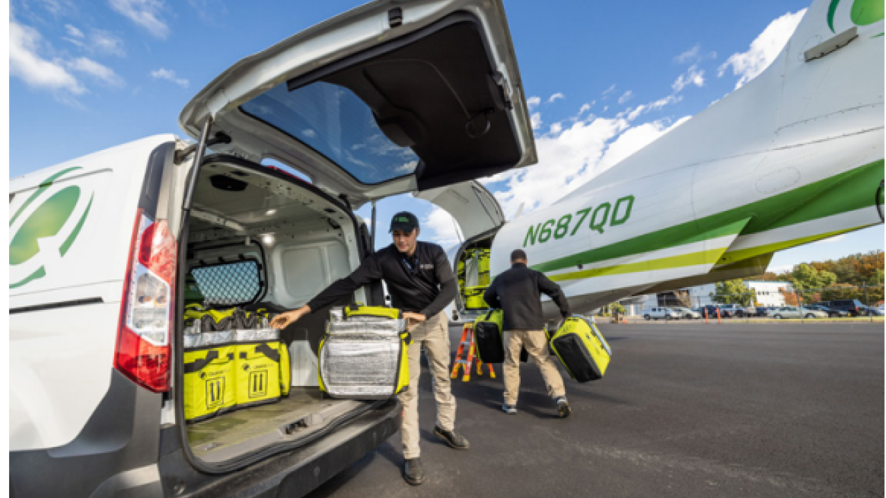 Two people unloading boxes from the back of a van, headed towards a small plane.