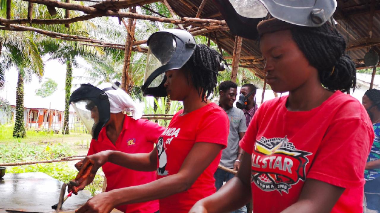 Three people each in a red shirt and welding masks, working at an outdoor table with welding tools and pieces of metal.