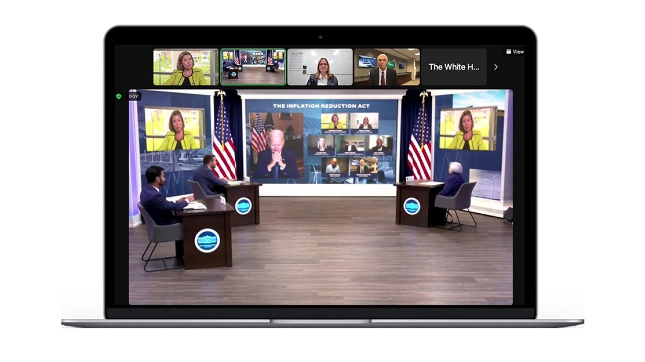three people at desks in an open studio, multiple screens behind them with government officials appearing virtually