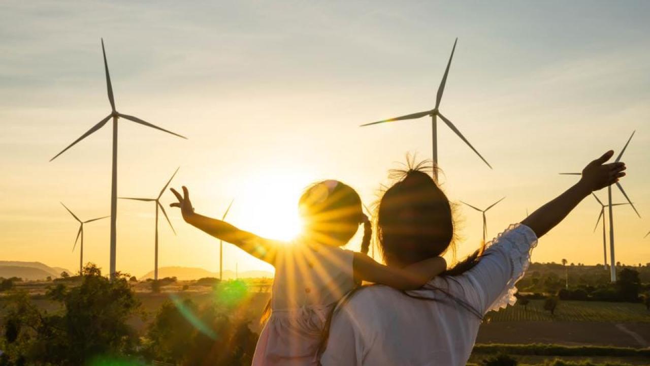 an adult holds a child and each have an arm stretched out. The sun is setting behind a filed of wind turbines