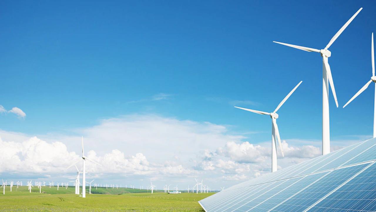 Outside on a bright day, windmills scattered on a large flat plain of grass, a solar panel in the right corner