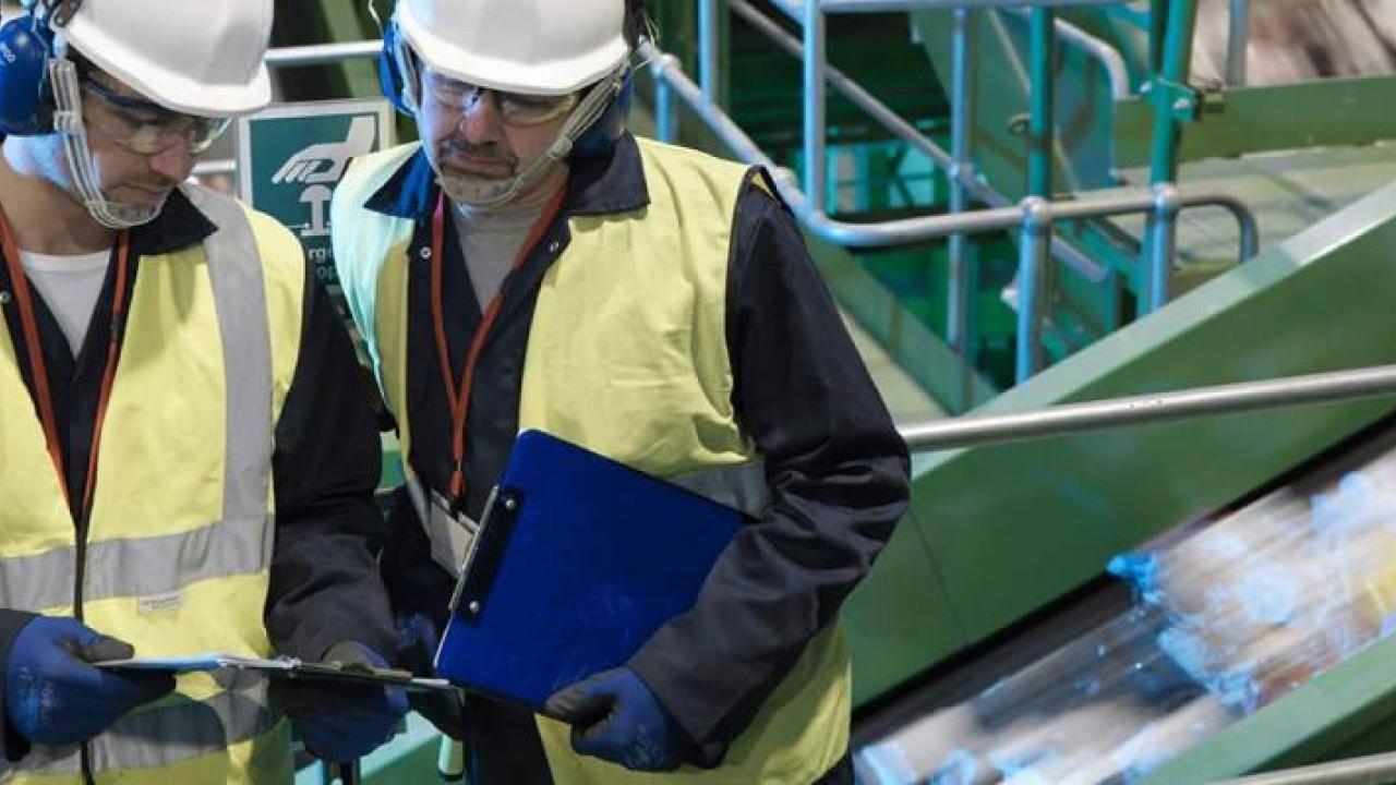 Two people in hard hats, protective wear and high-vis vests looking at a clip board. Fast moving items on a narrow ramp behind them.