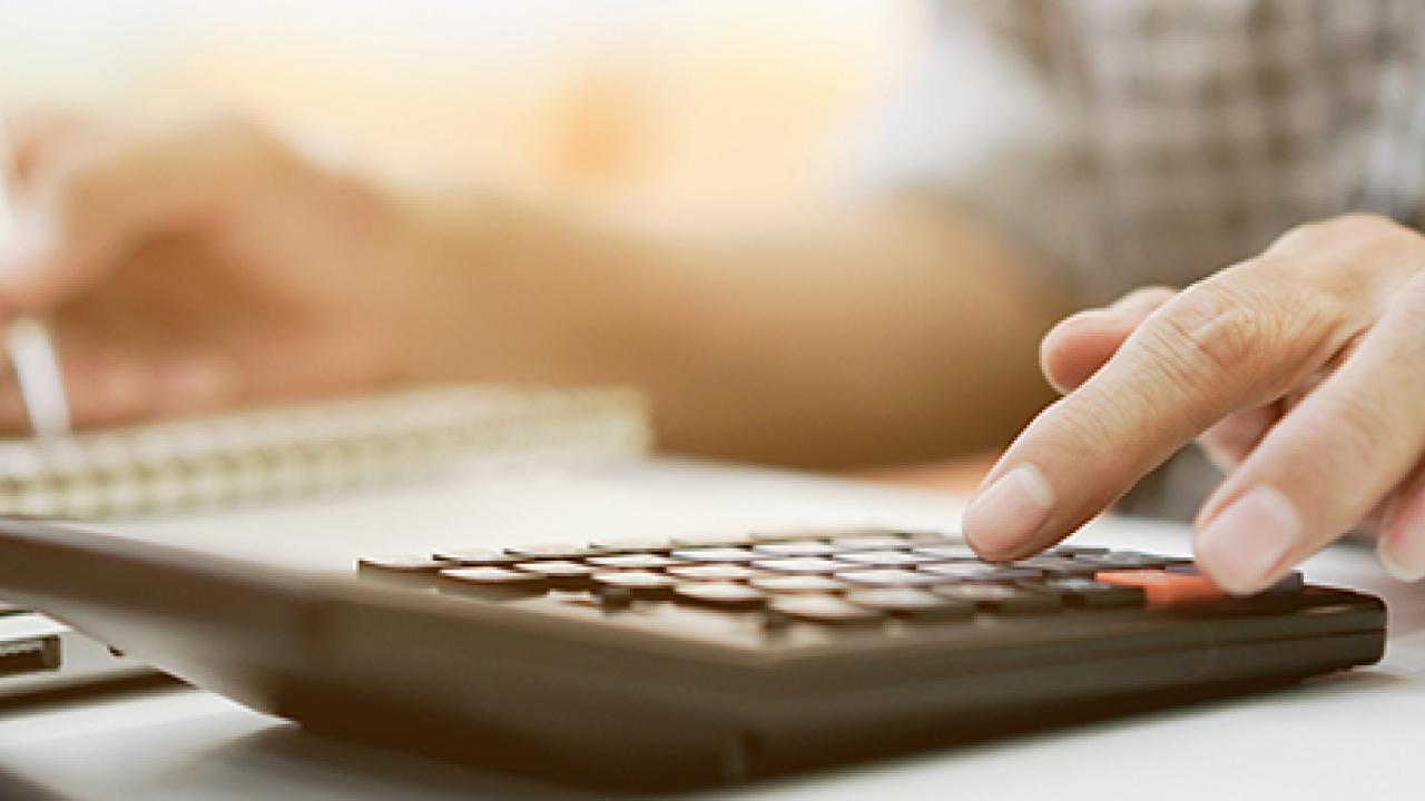 Close up of a person using a calculator, writing with the other hand and a laptop in front of them.