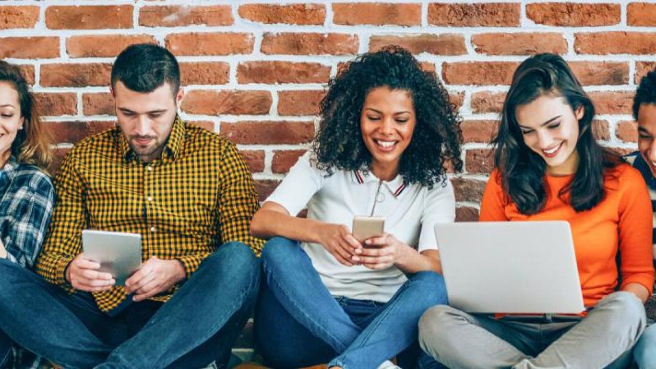 A group seated against a brick wall. Some smiling and looking at an open laptop or other devices.