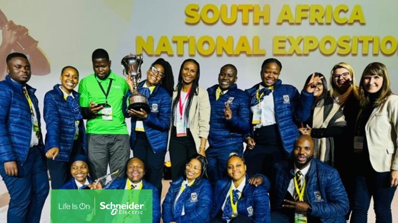 A group posed together, one holds a trophy and others a sign with Schneider Electric logo. "South Africa National Exposition sign behind them.