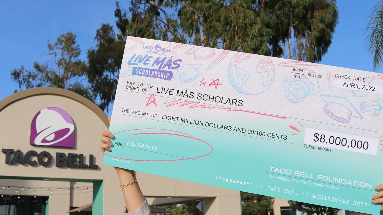A large check in front of a Taco Bell reads " Pay to the order of : Live Más Scholarships"