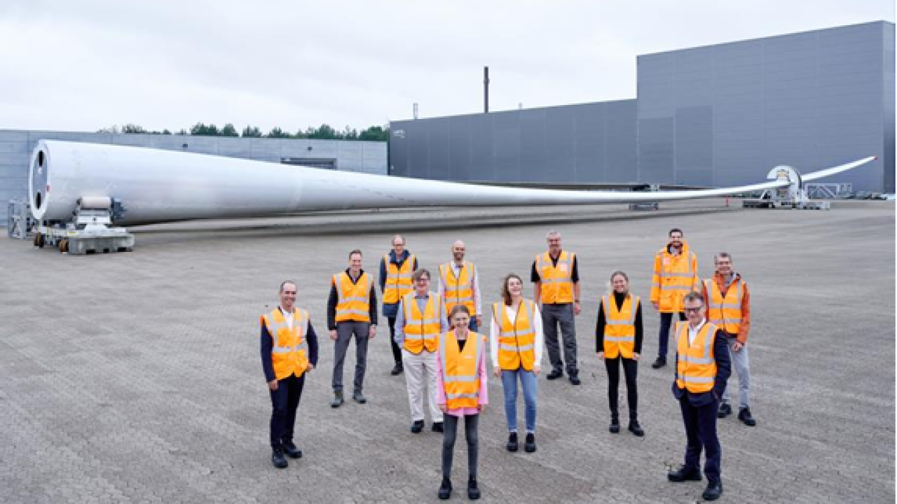 A group posed standing outside in front of a huge wind turbine blade.