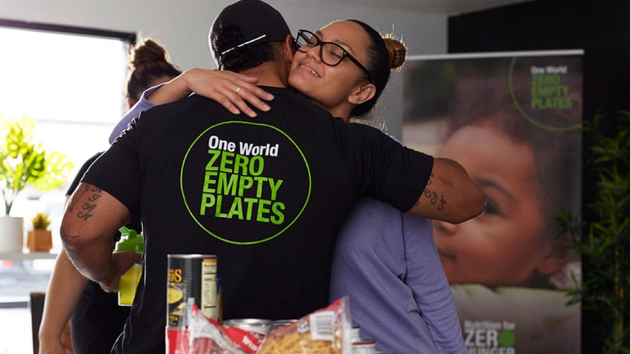 A person hugging someone with a t-shirt that reads: "One World, Zero Empty Plates"