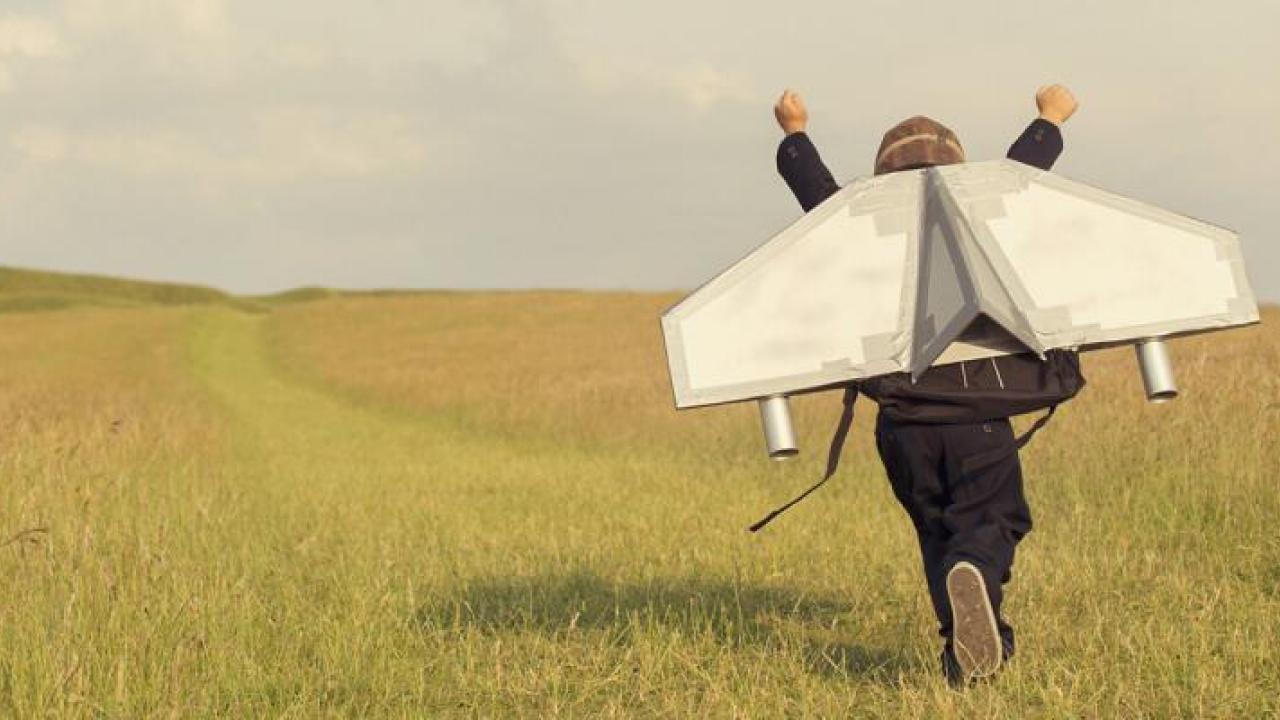 A child running in an open field with arms up and hand built wings attached to their back.