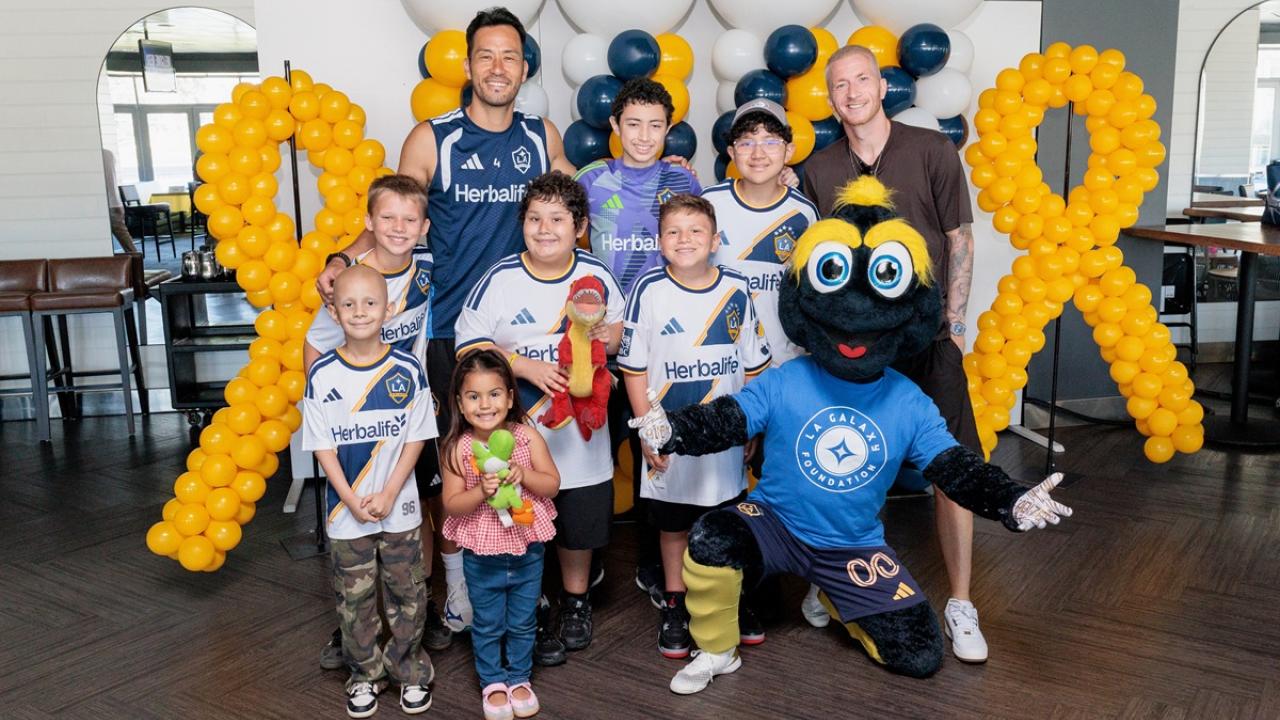 Youth participants, LA Galaxy players, and the LA Galaxy Foundation mascot Cozmo pose together in front of gold ribbon balloon displays during a community engagement event.