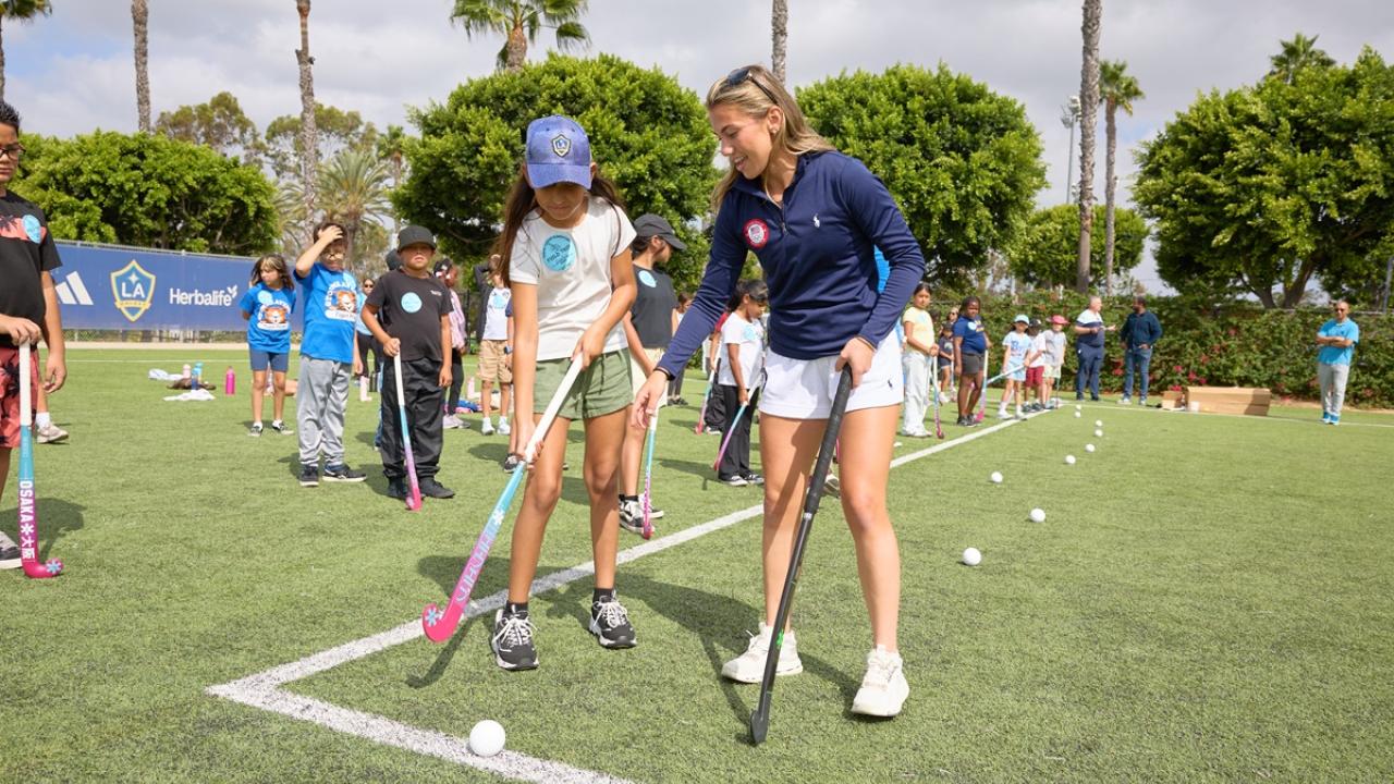 Ashley Sessa demonstrates field hockey stick positioning to a student as other students practice with sticks and balls on the field.