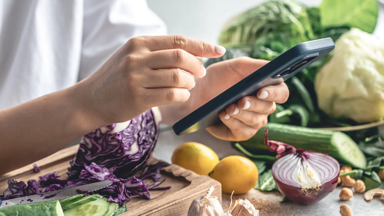 A person with a smart phone and fruits and vegetables on a counter.