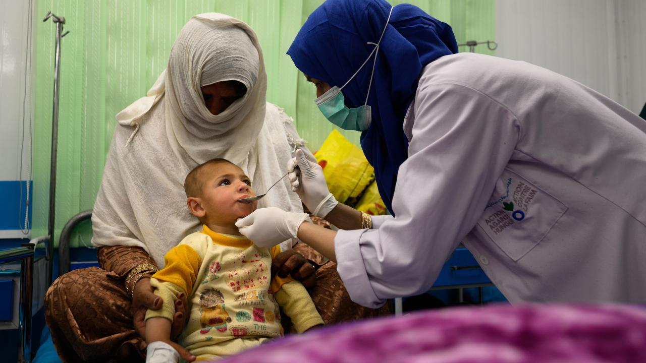 An ACF nurse leans in to give a spoonful of liquid medicine to a toddler. 