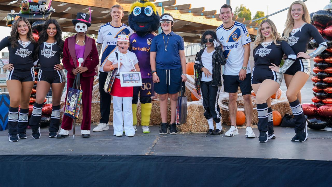 LA Kings Ice Crew members, LA Galaxy representatives, a costumed mascot and children in Halloween costumes pose together on a decorated outdoor stage.