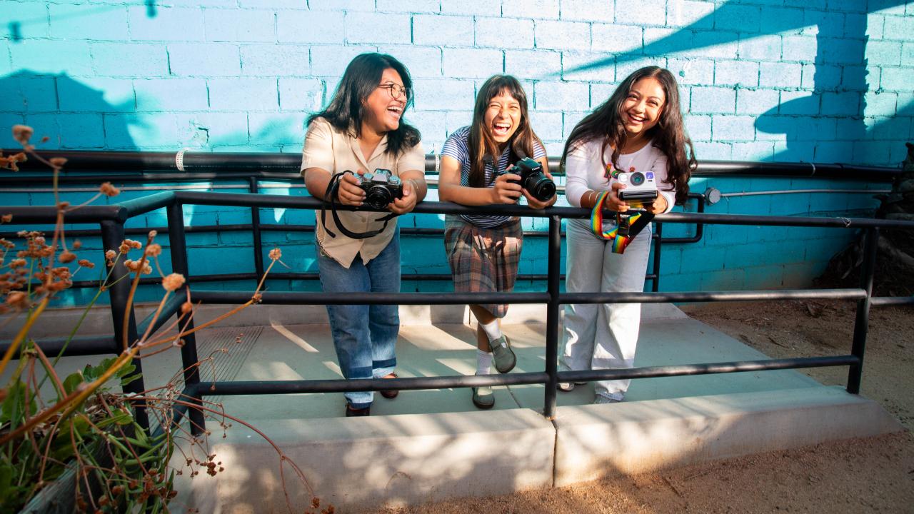 Three young people hold cameras and stand together along a railing in front of a bright blue brick wall.