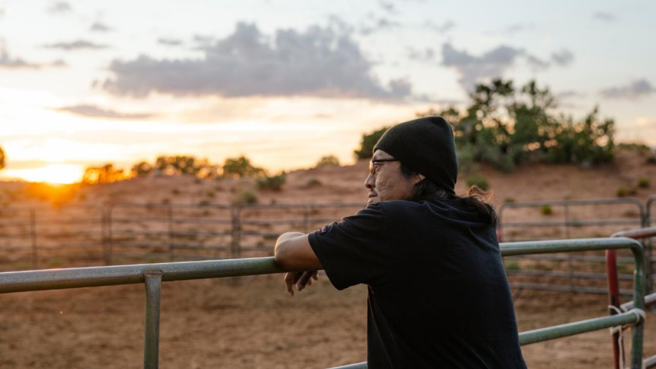 Nobert, a DaVita home dialysis patient, leans against a metal farm fence while he looks out at a sunset on Navajo Nation.