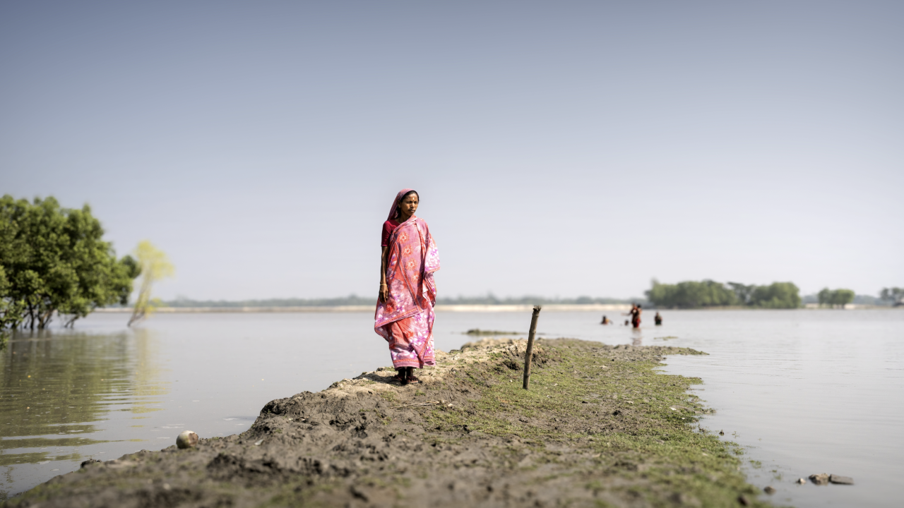 A woman stands on a narrow embankment surrounded by floodwater, with trees and people wading in the distance.