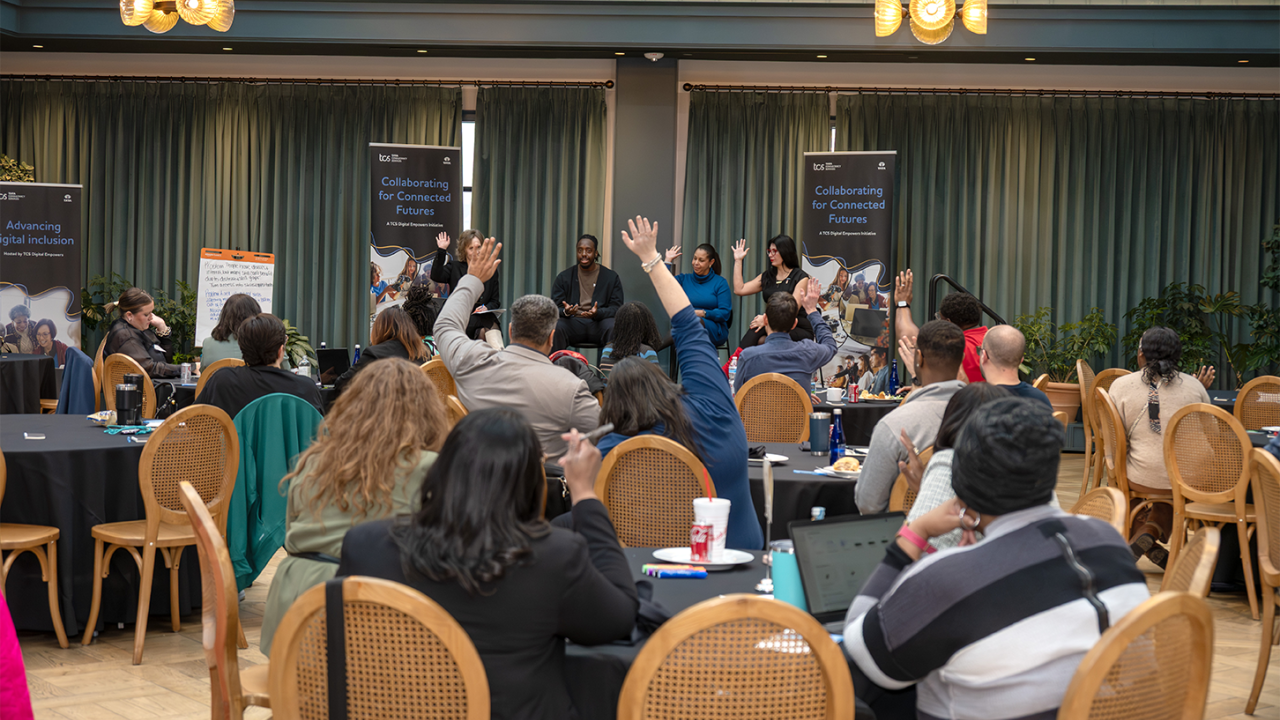 seated people raising hands at an event with speakers seated on stage