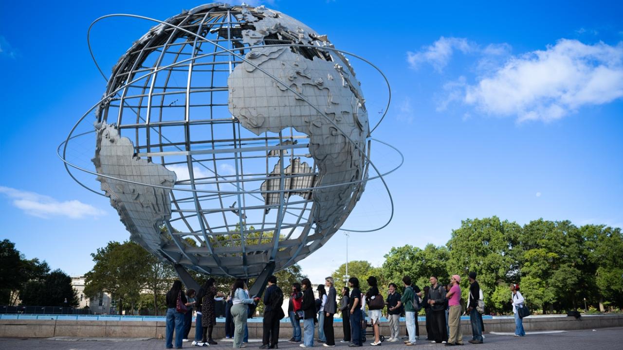 Group gathers outdoors beneath the Unisphere at Flushing Meadows Corona Park under a bright blue sky.