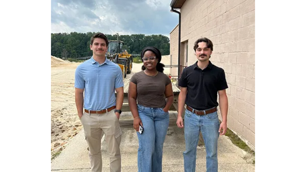The three trainees standing together next to a building, with a tractor and trees in the background